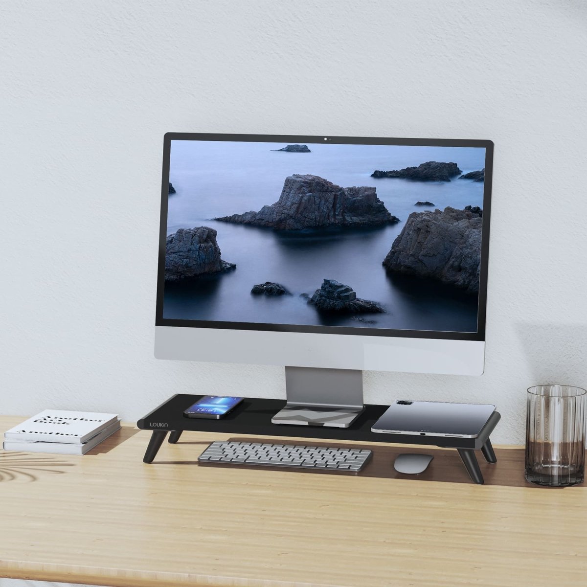 A stylish home office scene featuring the black Loukin Single Monitor Stand Riser (S4101). The stand elevates a large monitor to an ergonomic height and provides a storage space underneath for a keyboard and mouse. The stand's desktop holds a tablet and smartphone, demonstrating its organization and space-saving benefits on a wooden desk.