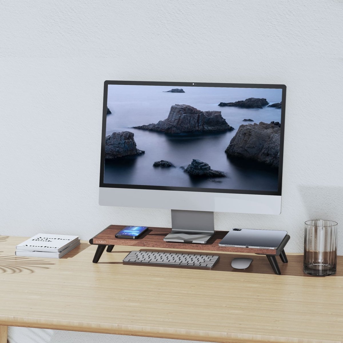 A home office scene featuring the Loukin Single Monitor Stand Riser (S4101) with a dark wood grain desktop and black legs. The stand elevates a large computer monitor, providing organized storage underneath for a keyboard and mouse. The stand's desktop holds a smartphone and tablet, optimizing desk space and promoting better ergonomics on a light wooden table.
