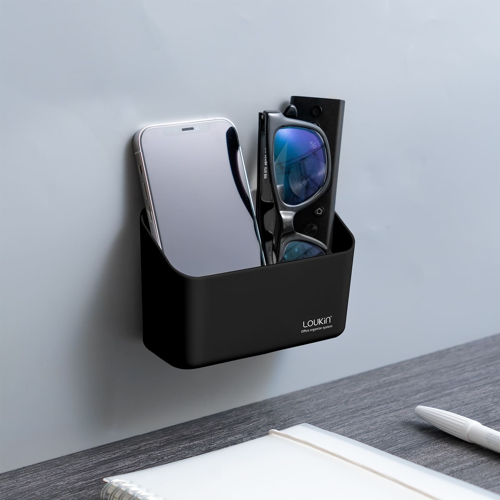 loukin W4101K Small Marker Holder in black, securely attached to a gray wall next to a desk. The compact organizer is being used to store a smartphone and a pair of glasses/remote control, demonstrating a small-scale office or personal item organization solution.