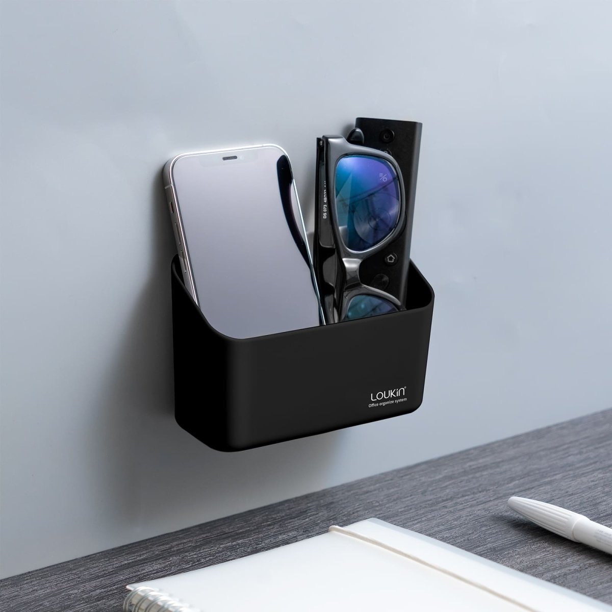 loukin W4101K Small Marker Holder in black, securely attached to a gray wall next to a desk. The compact organizer is being used to store a smartphone and a pair of glasses/remote control, demonstrating a small-scale office or personal item organization solution.