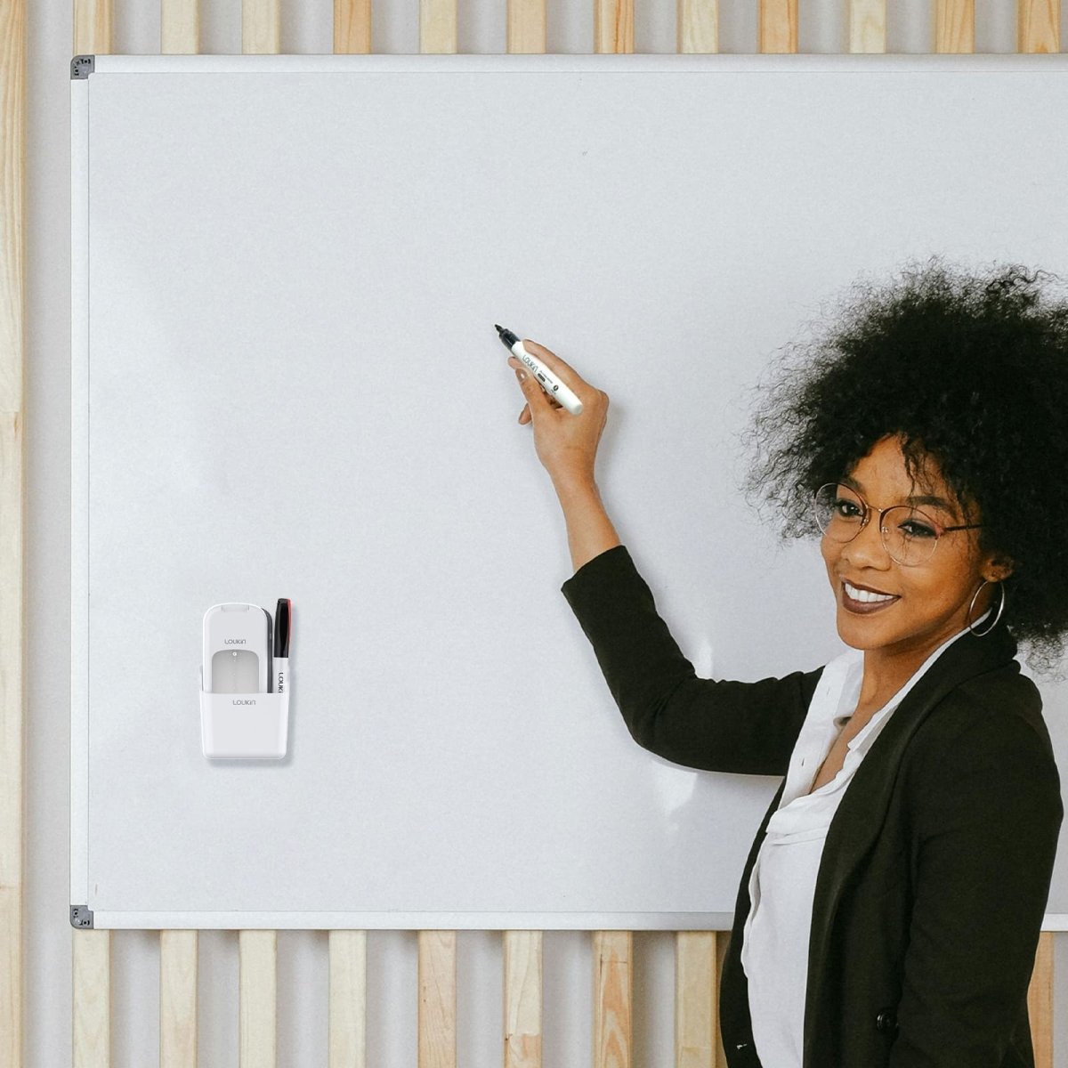 A professional woman is smiling and writing on a large, clean white magnetic whiteboard. The white loukin All-in-One Eraser (W3543K) accessory holder is magnetically attached to the left side of the board, securely holding a black dry-erase marker, showcasing organized accessory storage in an office or presentation setting.