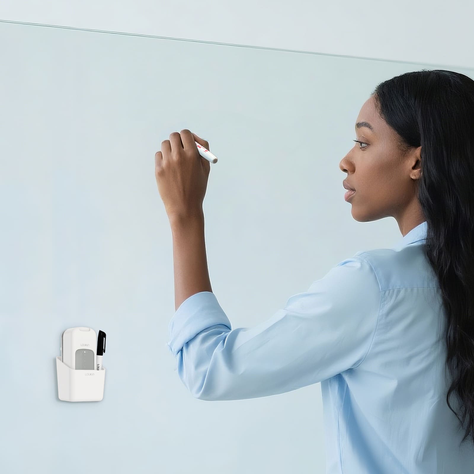 A professional woman in a light blue shirt is writing on a clear glass or white magnetic whiteboard. The white loukin All-in-One Eraser (W3543K) accessory holder is magnetically attached to the left side of the board, securely holding a black marker, showcasing convenient and tidy accessory storage in an office or presentation setting.