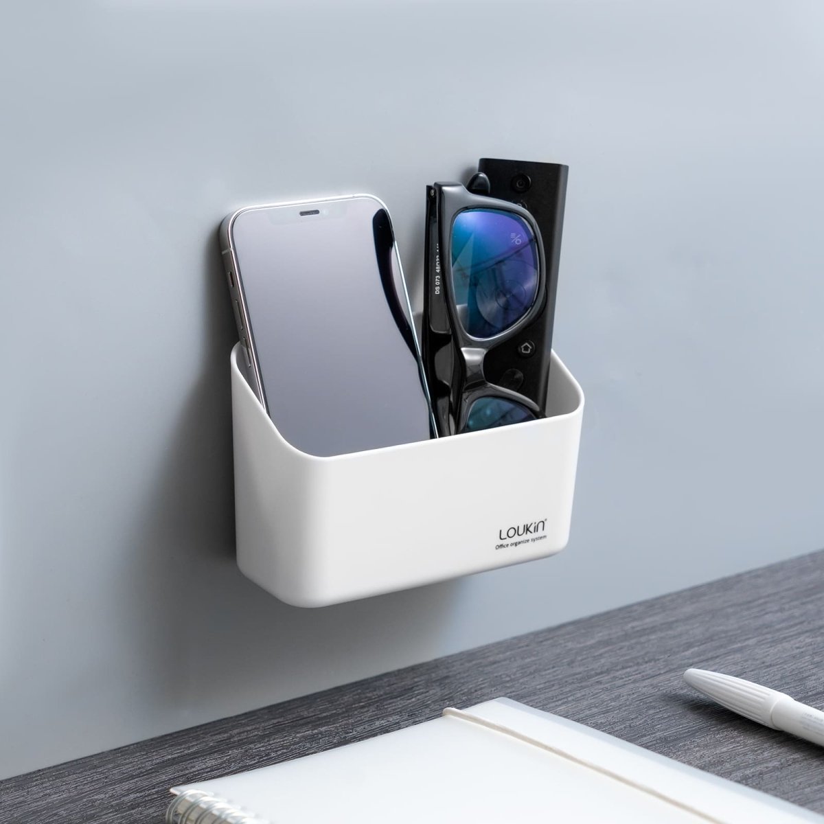 loukin W4101K Small Marker Holder in white, securely attached to a gray wall next to a desk. The compact organizer is being used to store a smartphone and a pair of glasses/remote control, demonstrating a small-scale office or personal item organization solution.
