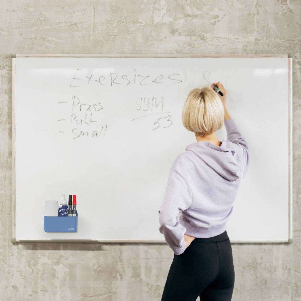loukin W4102 MarkerHolderSmall in blue/grey color, magnetically attached to a large white whiteboard in a fitness or workout setting. The holder is shown storing dry-erase accessories (markers and cleaner) while a blonde woman writes 'Exercises' and a workout plan on the board, demonstrating its use for organization in different environments.