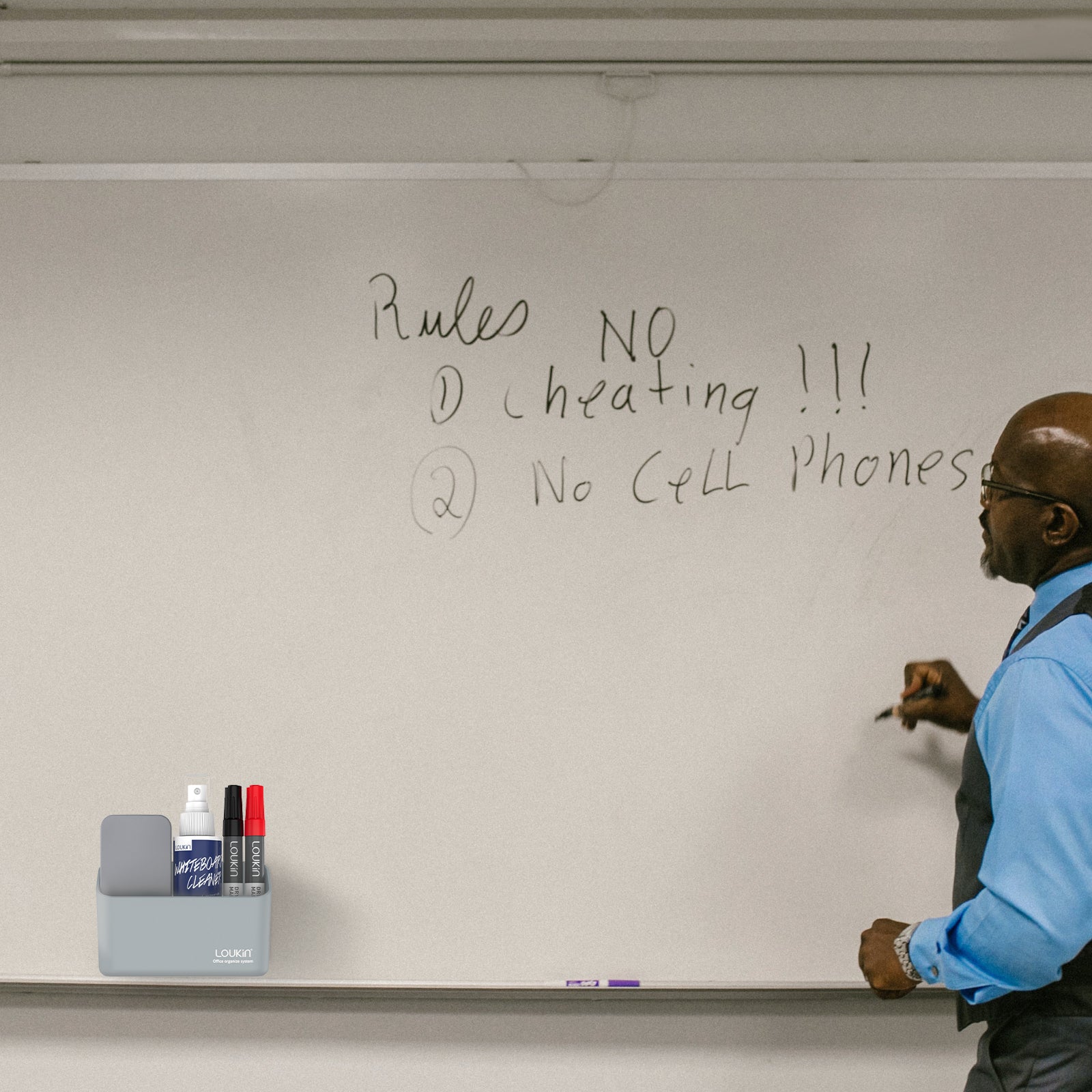loukin W4102 MarkerHolderSmall in grey, attached to a large white whiteboard in a classroom or office setting. The holder is shown storing an eraser, cleaner spray bottle, and markers (red and black) while a man in a blue shirt is writing on the whiteboard, demonstrating its use as a convenient magnetic accessory tray.