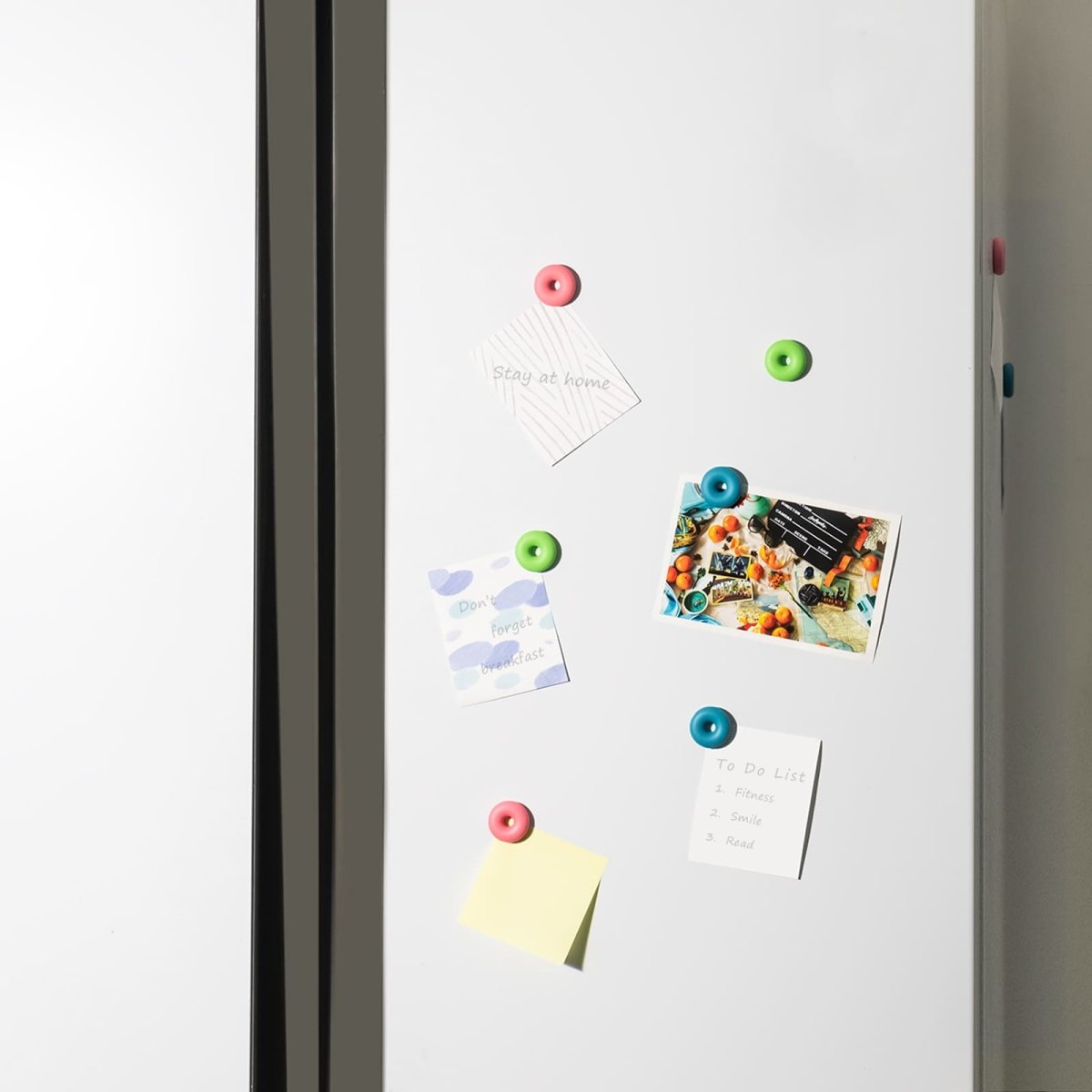 Use scenario of the colorful magnetic donut pushpins from the Loukin Big Marker Holder Set (W4102K). The magnets (blue, green, and pink) are shown holding up various notes, to-do lists, and photographs on a white magnetic surface, likely a whiteboard or refrigerator, emphasizing their practical application for organization and decoration.
