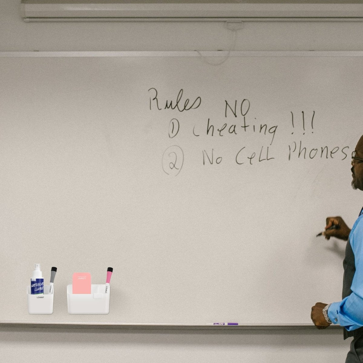 Lifestyle image featuring the Loukin Duo Magnetic & Adhesive Organizer Set (W4121K) in a classroom setting. A teacher is writing on a large whiteboard. The organizer set, consisting of a white marker/accessory caddy and a bottle of whiteboard cleaner, is placed on the bottom ledge of the whiteboard for easy access. The caddy holds dry-erase markers and a pink square eraser or sticky notes.